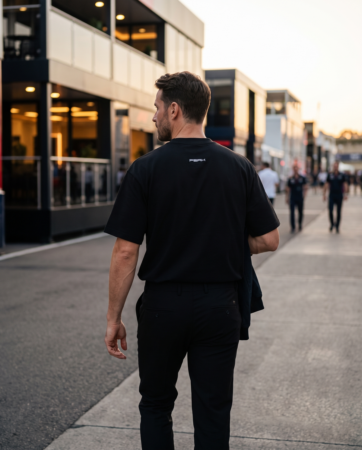 PEAK Back Neck Tee in Black, worn walking through a race paddock at dusk